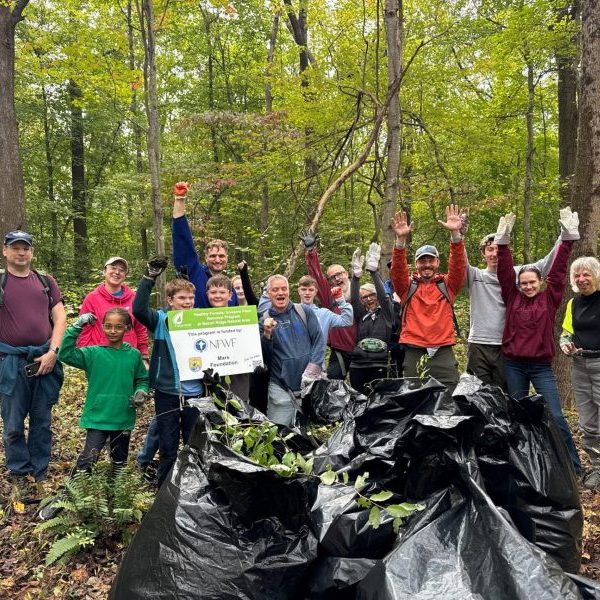 Large group of volunteers cheer and pose with "We Love Healthy Forests" Signs in front of a large pile of removed invasive plant species.
